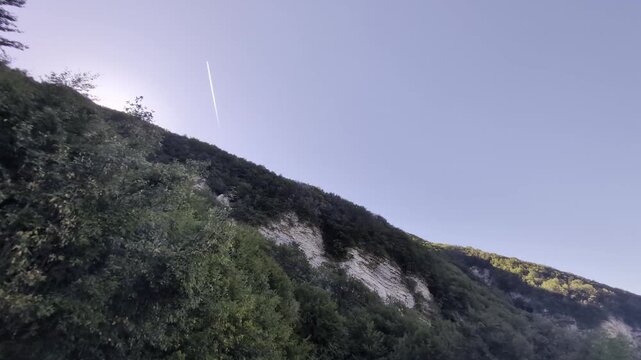 Inquadratura dal basso di un ripido costone montano alberato con pareti di roccia chiara affioranti, sovrastato da un cielo azzurro solcato da una scia aerea