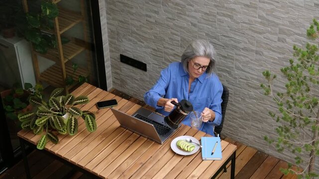 High angle view of a mature woman working remotely on her balcony and drinking coffee