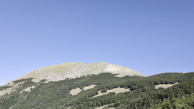 Suggestivo campo lungo di una cima montuosa brulla e rocciosa che si staglia contro un cielo azzurro e limpido, sovrastando una fitta fascia boscosa