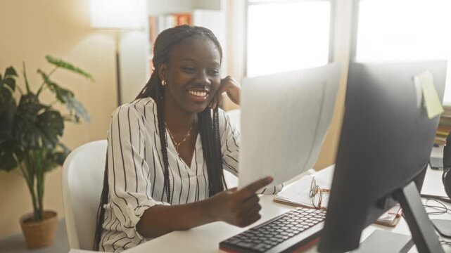 Woman smiles and points finger at a document while reading at a desk near a computer monitor and keyboard in an office building; confidence productivity.