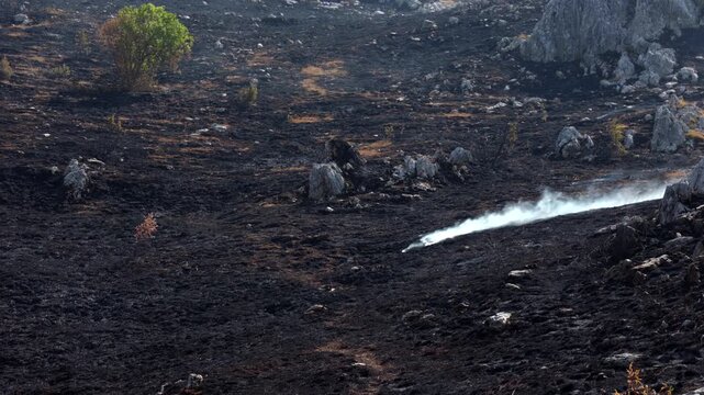 Smoke rises from smoldering ground after a wildfire, leaving a scorched rocky landscape with sparse surviving vegetation. Concept of natural disaster, environmental damage, climate change, wildfire.