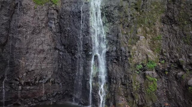Aerial pulling back from the rushing waters of Vaimahuta Waterfall as it splits over a dark, textured green basalt cliff face in Tahiti