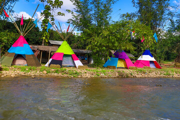 Row of vibrant teepee tents pitched along a flowing riverbank surrounded by green trees. © wannasak