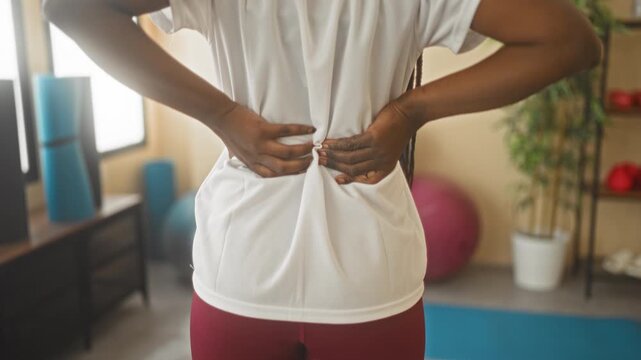 Woman holding lower back with both hands and gripping white shirt above red leggings in yoga studio; pain recovery.