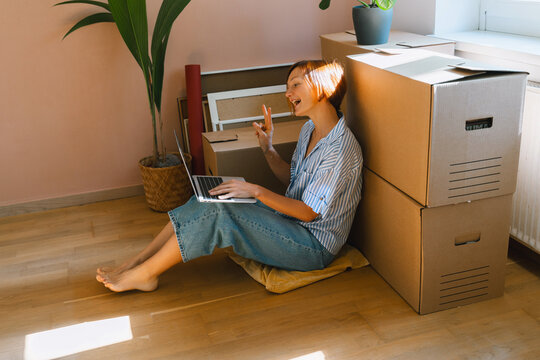 A smiling young woman sits on the floor with a laptop waving her hand next to moving boxes. Authentic first home ownership and real estate property investment concept.