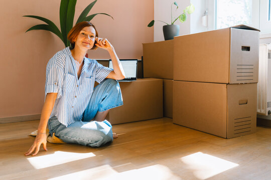 A smiling young woman sits on the wooden floor looking away from her laptop during a move. Authentic first home ownership and real estate property investment concept.