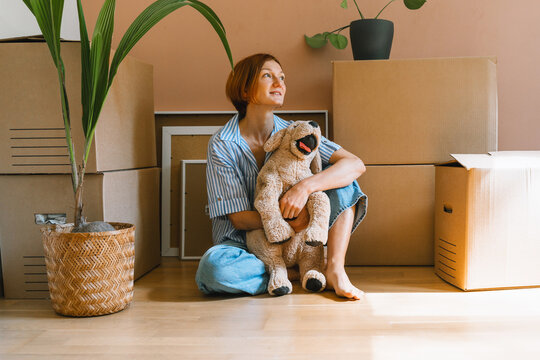 A happy young woman sits on the floor with plush dog toy next to a potted houseplant. Authentic first home ownership and real estate property investment concept.