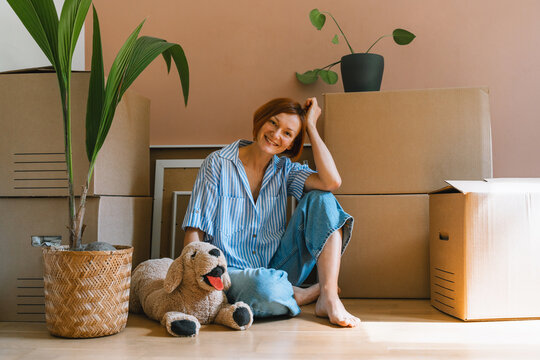 A happy young woman sits on the floor with plush dog toy next to a potted houseplant. Authentic first home ownership and real estate property investment concept.