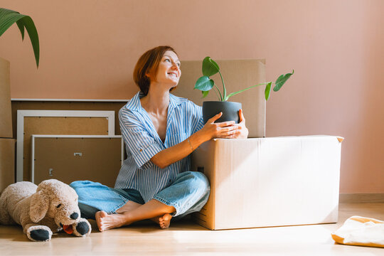 A happy young woman sits on the floor with plush dog toy next to a potted houseplant. Authentic first home ownership and real estate property investment concept.