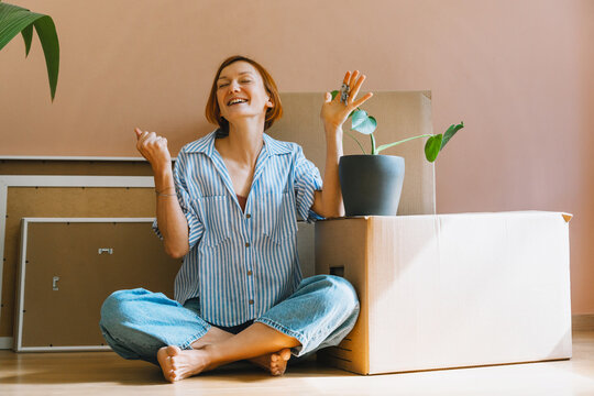 A laughing young woman holds up her house keys while sitting cross-legged among moving boxes. Authentic first home ownership and renting property investment concept.