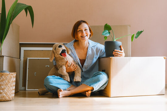 A happy young woman sits on the floor with plush dog toy next to a potted houseplant. Authentic first home ownership and real estate property investment concept.