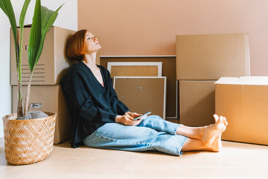A thoughtful young woman in a black shirt sits on the floor looking up among moving boxes. Authentic first home ownership and real estate property investment concept.
