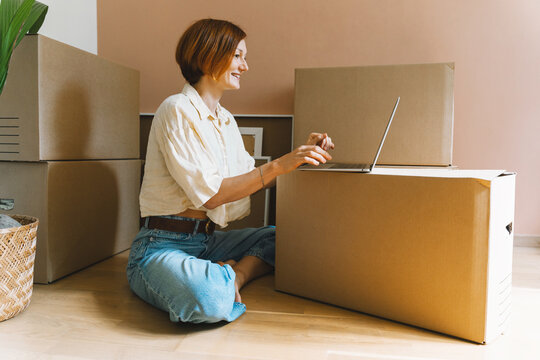 A young woman types on her laptop placed on a cardboard box in an empty sunny room. Authentic first home ownership and real estate renting property concept.