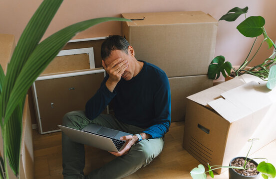 A frustrated man holds his head while sitting on the floor with a laptop surrounded by boxes. Authentic home relocation and real estate mortgage pressure concept.