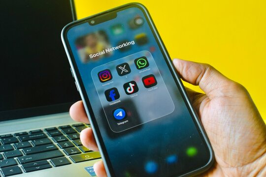 Sarawak, Malaysia - 8 August 2026: Person holding a smartphone displaying popular social networking applications in front of an open laptop on a vibrant yellow background.