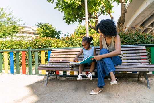African american mother and young daughter bonding outdoors, reading a book together on a park bench. Enjoying summer day