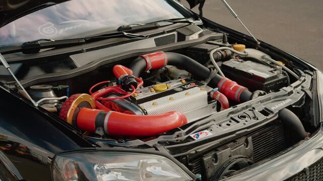 At a car festival, a modified car engine is on display. The engine features bright hoses and various components. Enthusiasts gather to appreciate the work put into it.