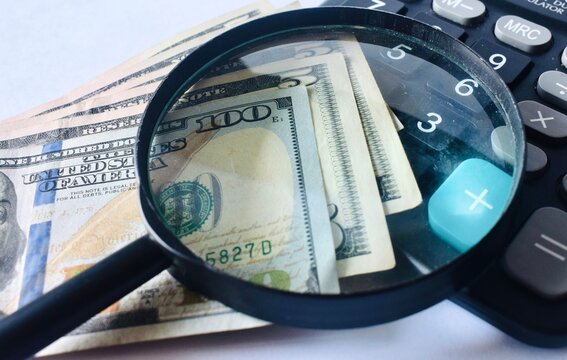 Close-up of US dollar bills under a large magnifying glass positioned over a desktop calculator, conceptualizing financial audit, tax review, and budget analysis.