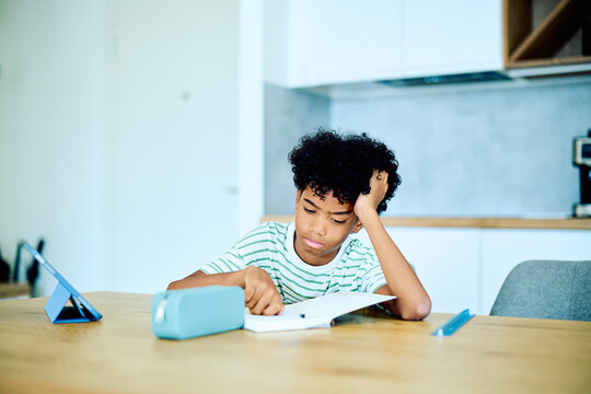 Young african american boy sitting at table, learning and feeling tired or frustrated while doing homework with a tablet nearby