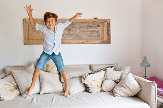 Happy, child or jumping on sofa for holiday, fun weekend or summer break in living room or home. Portrait, kid or playful boy with smile in air on couch for childhood vacation or celebration in house