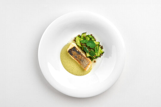 Gourmet fried fish fillet with green sauce, zucchini ribbons, and microgreens in a white bowl, isolated on a white background.