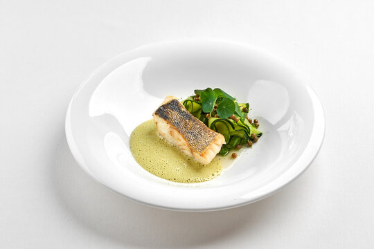 Gourmet fried fish fillet with green sauce, zucchini ribbons, and microgreens in a white bowl, isolated on a white background.