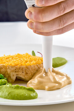 Chef applying sauce with a piping bag onto a gourmet fish dish with green puree in a white plate. High-end dining prep.