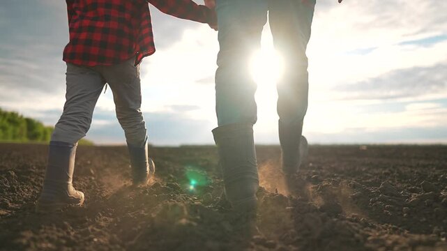 Father and child walking hand in hand through empty field at sunset. Sky glows warmly above their heads. Child walks beside father in silence. Outdoor stillness wraps father and child together