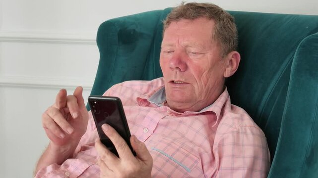 Senior man relaxes in comfortable armchair, focused on interacting with his smartphone