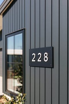 Black metal house number plaque 228 mounted on charcoal vertical siding beside a steel-frame window