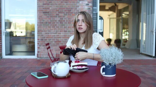 Woman opens her wallet at an outdoor cafe and looks shocked after realizing there is no money left to pay for cake and tea creating an awkward unexpected situation at all