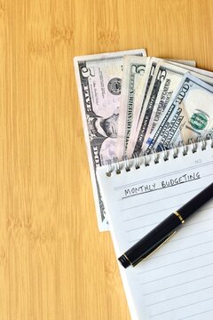 Top view flat lay of US dollar bills tucked under a notebook with the title "MONTHLY BUDGETING" and a black pen on a wooden background.