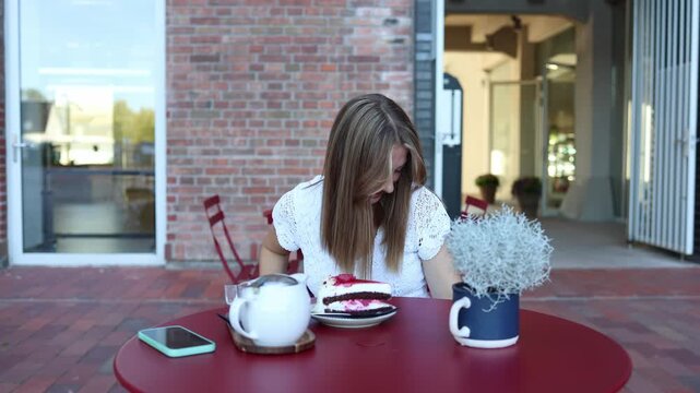 Woman notices something has fallen under the cafe table and reaches down to pick it up while sitting beside cake and tea during a casual afternoon break at outdoor cafe