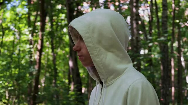 Teenage boy wearing white hoodie standing in sunny green forest, looking around and smiling outdoors