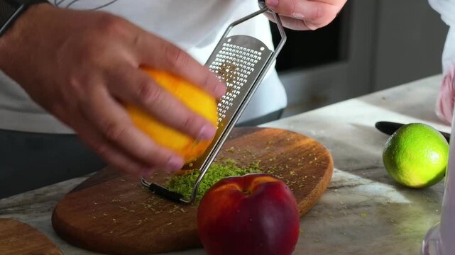 Close up of person grating fresh orange peel with metal tool on wooden cutting board in kitchen
