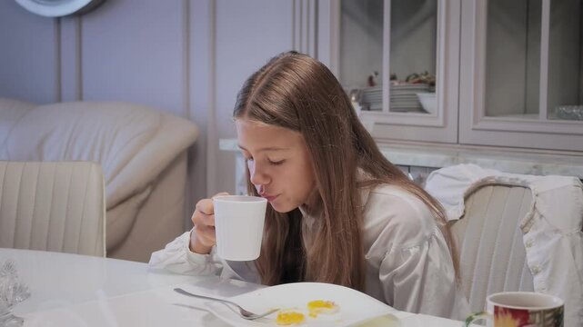 Young girl drinking from cup at table with breakfast, indoor setting
