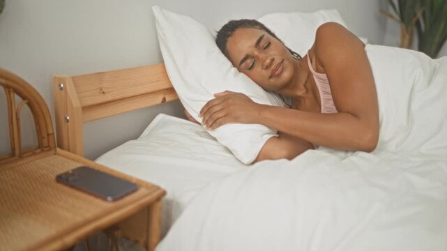 Woman lying in bed reaching with bare arm to grab a smartphone from a bedside table in a bright studio bedroom set; serene morning.
