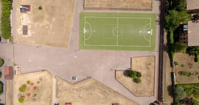 Aerial top view of a football pitch and adjacent dirt sports areas in a park, bordered by trees, houses, and a road. Zenithal shot.