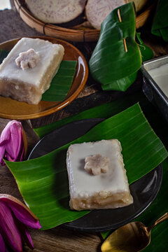 Traditional Thai tako taro pudding dessert square cut topped with thick coconut milk cream in aluminum tray next to banana leaf parcels and fresh sliced taro root. Asian sweet food snack.