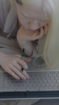 Slow motion top view of albino lady studying online typing with laptop and writing in notebook lying in bed at home. Distance education and student concept.