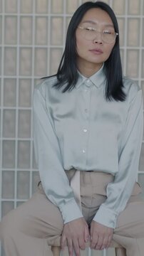 Slow motion portrait of beautiful Asian lady in elegant outfit sitting indoors against stylish interior. Confident person and luxury apartment concept.
