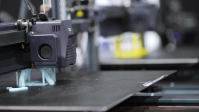 3D printer extrudes blue filament onto black build plate. Printer head moves precisely. Background contains blurred tools and materials