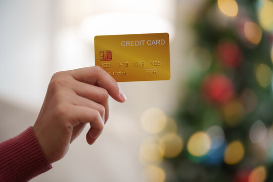 Close-up of a hand holding a credit card with a festive Christmas tree in the background, representing holiday shopping, seasonal spending, cashless payment, retail sales and discounts.
