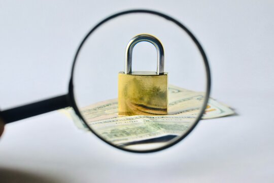 A magnifying glass zooming in on a brass padlock locked over US dollar bills, representing financial audit, security inspection, and fraud prevention analysis.