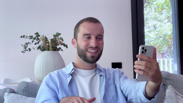 Young man sitting on a couch at home, smiling and gesturing while talking with friends or family on a video call, using his modern smartphone for communication and social distancing