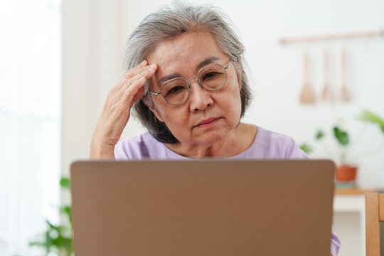 Close up of stressed, tired senior Asian woman with gray hair and eyeglasses working, looking at computer laptop, put her hand on forehead in confusion in bright cozy, modern living room at home