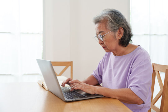 Senior Asian woman with gray hair and eyeglasses sitting at wooden table, working, typing on computer laptop in bright cozy, modern living room at home with concentration. Active lifestyle, technology