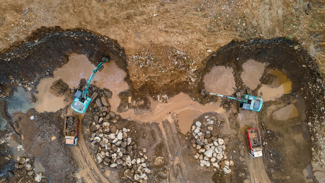 Excavator and Dump Truck Working in a Large-Scale Rock and Sand Quarry Near a Rocky Cliff. Heavy Machinery Operates at Mining Site with Raw Materials and Rugged Terrain view from above.