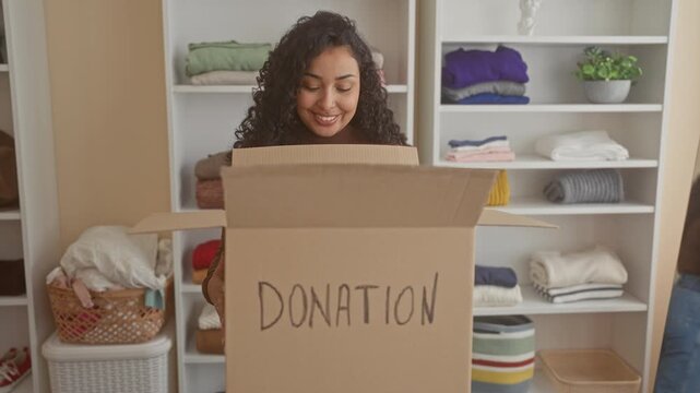 Woman holding donation box with folded clothes on shelves inside a donation sorting building; compassion generosity care.