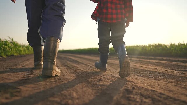 Boy follows farmer down quiet road. Boots stir light dust. Walking side by side under warm sunset. Field stretches calm. Summer road holds footprints. Man keeps watch on happy child enjoying path.